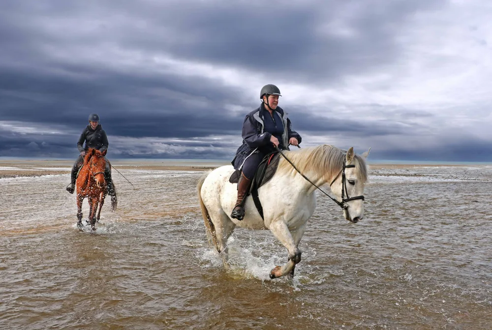 Two riders in shallow water on a beach under dark clouds